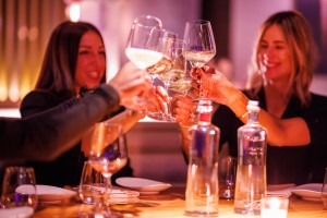 A close up of people holding up wine glasses in cheers, woman smiles in background.