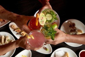 A close up from above of people holding cocktails in cheers, various plates of food slightly visible below.
