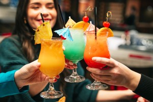 A close up of three people holding colorful cocktails in cheers, a woman smiles in the background.