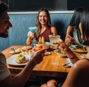 A group of friends seated around a wooden table, toasting with colorful cocktails. A guest is sitting on a blue upholstered banquette, smiling and holding a blue cocktail. The table is filled with a variety of sharing plates, including gyoza, rice topped with a fried egg, salad, and hibachi-fried dishes.